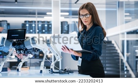 Portrait of a Young Robotics Engineer Using Laptop Computer, Analyzing Robotic Machine Concept in a High Tech Factory. Female Scientist Manipulate and Program the Robot for Work.
