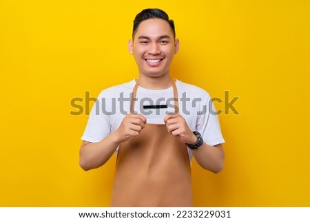 successful smiling young Asian man 20s barista employee wearing brown apron working in coffee shop, holding credit bank card, looking at camera on yellow background. Small business startup concept