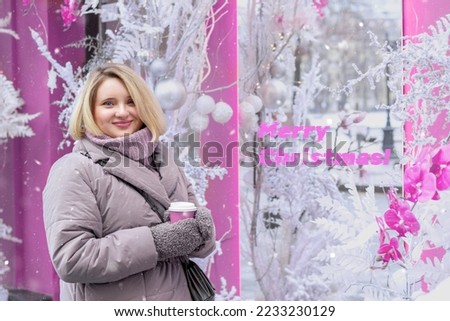 A young woman in warm clothes in a snowy city in winter enjoys coffee and wishes Merry Christmas. Christmas decor. In pink tones.