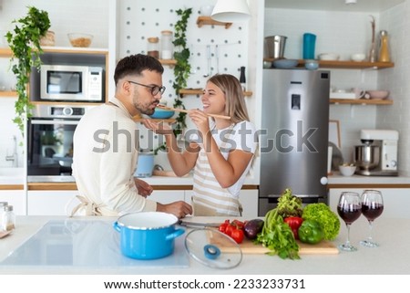 Beautiful young couple is feeding each other and smiling while cooking in kitchen at home. Happy couple is preparing healthy food on light kitchen. Healthy food concept. Royalty-Free Stock Photo #2233233731