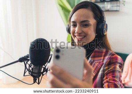 Latin american young woman wearing headphones standing by professional microphone smiling looking at smartphone recording herself. Influencer singer. Horizontal indoor shot. High quality photo