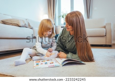 Smiling young mother sit on warm floor play with little infant toddler child, happy biracial mom relax have fun read book with small baby girl at home, motherhood, childcare concept
