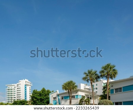 Views of white buildings with trees outdoors under the clear blue sky at Miami, Florida. There are small buildings at the front and a view of tall building with balconies on the left at the back.