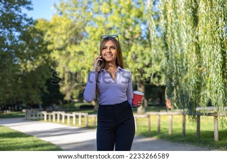 Woman with brown hair walks in the park and has a phone call with her smartphone. She holds a coffee cup. Outdoor, public park, relaxed position, leisure activity, summer and casual clothing. 