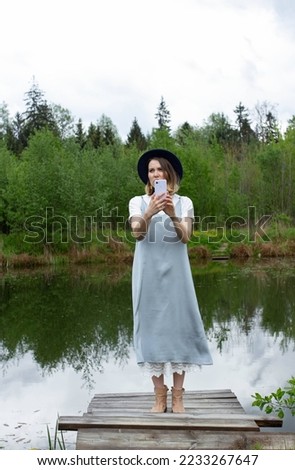Happy hipster young woman in blue dress and hat outdoor holding mobile phone, blogger girl taking pictures by camera phone, person with smartphone near the lake and forest, full length photo