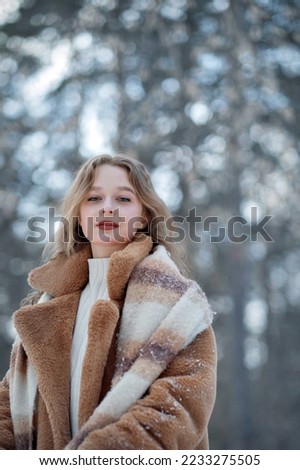 A white blonde girl in a knitted hat, a long scarf and a fur coat walks through the forest and smiles. Woman catches snow in white mittens. Winter is on the street. High quality photo. Christmas Eve