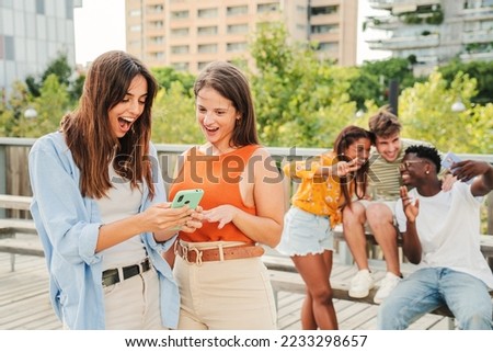 Two attractive and cheerful smiling women looking and showing the last funny and entertaining news on their smartphone outside at the university campus. At background, three young friends, having fun