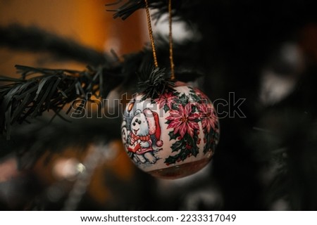 Close-up of a painted Christmas bauble decorated with joyful poinsettias and cartoons hanging on tree branch with warm yellow background