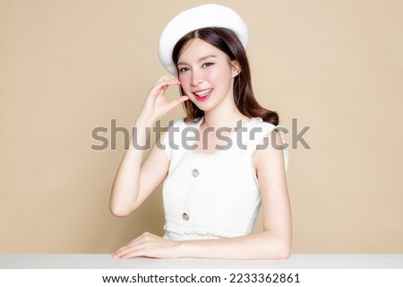 Cute Asian woman with perfect skin and show love Sign. Pretty girl model wearing white beret and natural makeup on beige background. Cosmetology, beauty and spa, wellness, Plastic surgery.