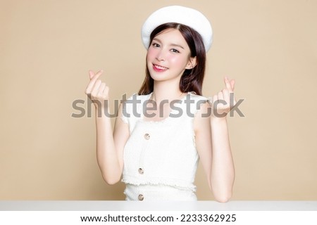 Cute Asian woman with perfect skin and show love Sign. Pretty girl model wearing white beret and natural makeup on beige background. Cosmetology, beauty and spa, wellness, Plastic surgery.