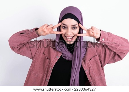 Cheerful positive young beautiful woman wearing hijab and pink overshirt over white background shows v-sign near eyes open mouth