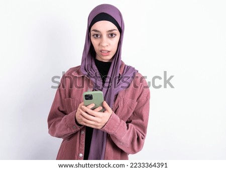 Photo of astonished crazy young beautiful woman wearing hijab and pink overshirt over white background hold smartphone dislike feedback concept
