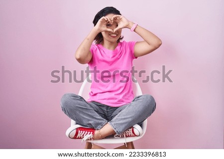 Hispanic young woman sitting on chair over pink background doing heart shape with hand and fingers smiling looking through sign 