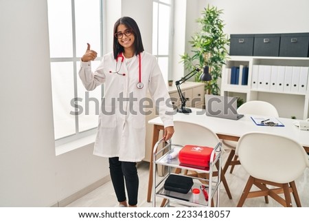 Young hispanic woman wearing doctor uniform and stethoscope smiling happy and positive, thumb up doing excellent and approval sign 