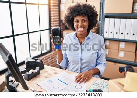 Black woman with curly hair working at small business ecommerce holding credit card and dataphone looking positive and happy standing and smiling with a confident smile showing teeth 