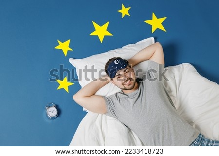 Top view calm young smiling man in pajamas jam sleep mask rest relax at home lies in bed hold hands behind head neck look camera isolated on dark blue sky background Good mood night bedtime concept