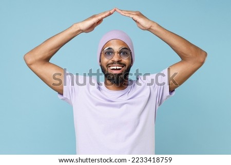 Young happy positive man of African American ethnicity he wear purple t-shirt glasses hold folded hands above head like house roof stay home isolated on plain pastel light blue cyan color background