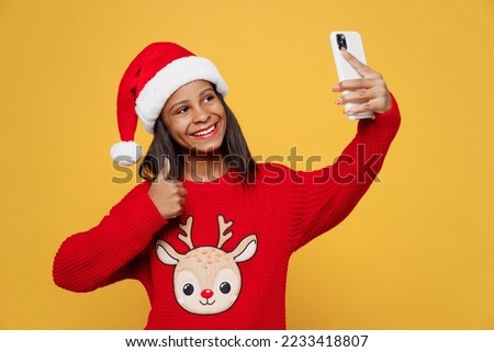 Merry little kid teen girl 13-14 years old wear red xmas sweater with deer Santa hat posing do selfie shot on mobile cell phone isolated on plain yellow background. Happy New Year 2023 holiday concept