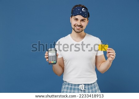 Young rich man in pajama jam sleep mask rest at home holding wireless bank payment terminal to process and acquire credit card payments isolated on dark blue background Good mood night bedtime concept