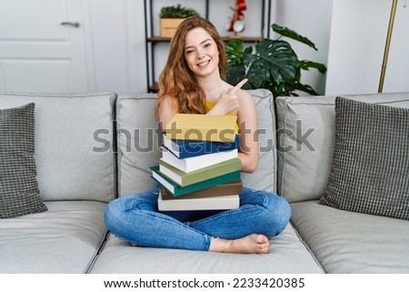 Young caucasian woman sitting on the sofa with books at home with a big smile on face, pointing with hand and finger to the side looking at the camera. 