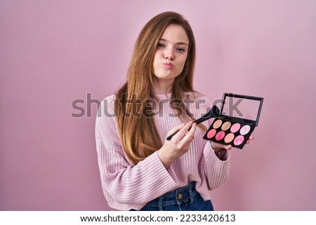 Young caucasian woman holding makeup brush and blush palette puffing cheeks with funny face. mouth inflated with air, catching air. 