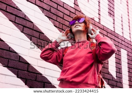 Stylish woman in magenta color jacket and heart shaped sunglasses wearing wireless headphones on her head and listening music. Urban city street fashion. Color of the 2023 year. Selective focus. Royalty-Free Stock Photo #2233424415