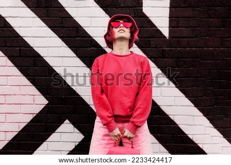 Hipster fashion young woman in trendy magenta color sweatshirt and sunglasses and bucket hat posing on the painted brick wall background. Color of the 2023 year. Urban city street fashion. Royalty-Free Stock Photo #2233424417