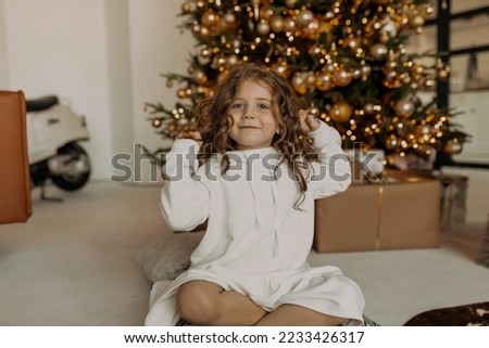Happy charming cute little girl with wavy hair wearing white dress resting at home in front of Christmas tree. 