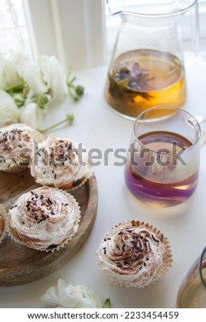 Muffins with cream and chocolate sprinkles on a wooden stand near white ranunculus and a cup of tea