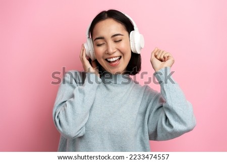 Happy satisfied girl standing listening favourite music with headphones, closed eyes and toothy smiling, Indoor studio shot, isolated on pink background  Royalty-Free Stock Photo #2233474557