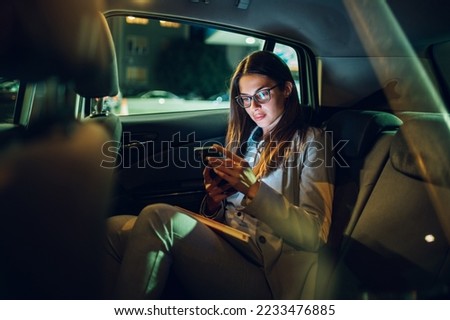 Beautiful young businesswoman traveling with car on a business trip during night while sitting in a back seat and using a smartphone. Gorgeous female using mobile phone to send email or messages. Royalty-Free Stock Photo #2233476885