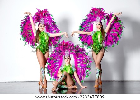 Three young brunette girls dancers in carnival costumes of green and pink feathers pose on a white background