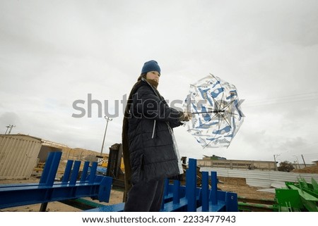 cool stylish boy in a coat and hat with a scarf posing plays and walks through pipes with an umbrella in an industrial area among green yellow blue metal structures against a cloudy sky on a rainy day