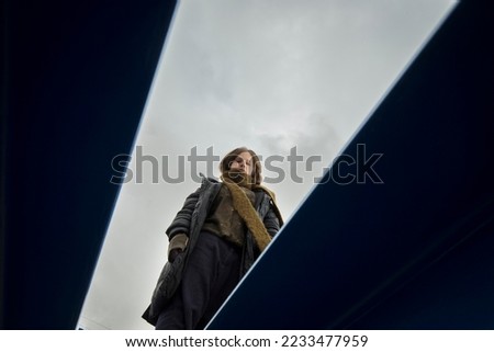 cool stylish caucasian boy in a coat and hat with a scarf posing coolly plays and walks through pipes in an industrial area among green yellow blue metal structures against a cloudy sky on a rainy day