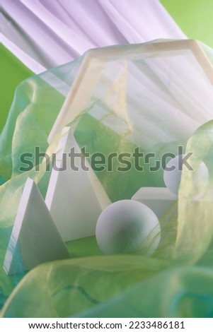 White geometric figures still life composition. Prism pyramid sphere objects beyond transparent green fabric. Vertical photo.