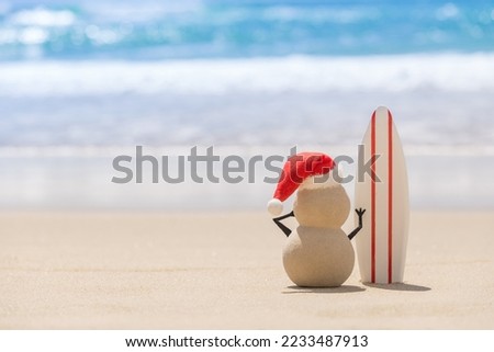 Sandy Christmas Snowman is watching the waves, standing on beautiful beach with a surf board - horizontal version Royalty-Free Stock Photo #2233487913