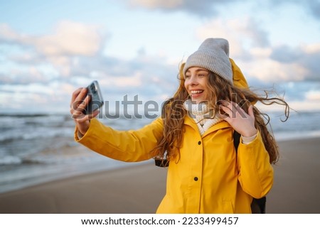 Young tourist with phone. Selfie time. Tourist in a yellow jacket posing by the sea at sunset. Travelling, lifestyle, adventure. Royalty-Free Stock Photo #2233499457