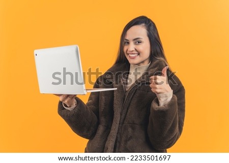 Young adult latin american woman wearing brown fur coat holding up laptop and looking to camera with wide smile and showing thumbs up gesture. Advertising. Online shopping, Yellow background