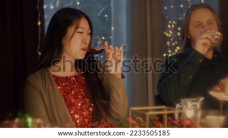 Friends clink glasses, drinking wine and talking. Multi cultural friends celebrating Christmas or New Year 2023. Served holiday table. Warm atmosphere of Christmas dinner at home. Christmas party.