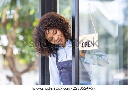 Happy waitress standing at restaurant entrance. Portrait of african american business woman attend new customers in her coffee shop. Happy woman owner showing open sign in her small business shop.