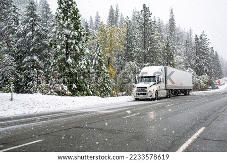 White big rig industrial semi truck with grille guard transporting cargo in dry van semi trailer standing on road shoulder of a winter highway during a snow storm near Shasta Lake in California Royalty-Free Stock Photo #2233578619