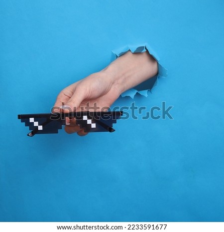 Female hand holds 8 bit pixel sunglasses through torn hole in blue paper