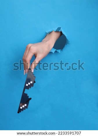 Female hand holds 8 bit pixel sunglasses through torn hole in blue paper