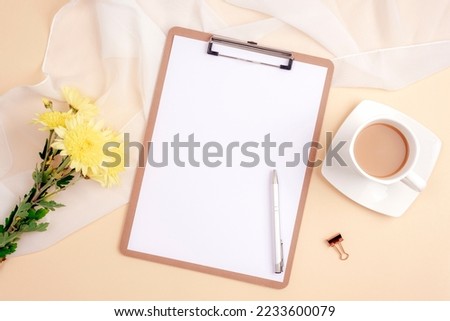 Desktop with blank clipboard, pen, coffee cup and yellow chrysanthemum flowers on light beige background. Top view, flat lay, mockup.