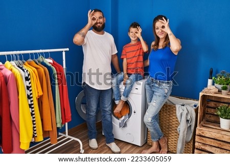 Family of three at laundry room smiling happy doing ok sign with hand on eye looking through fingers 