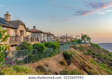 Two-storey houses near the edge of a slope at San Diego, California. There is an iron fence near the shrubs at the front of the two-storey houses and a view of a sunset sky at the background.