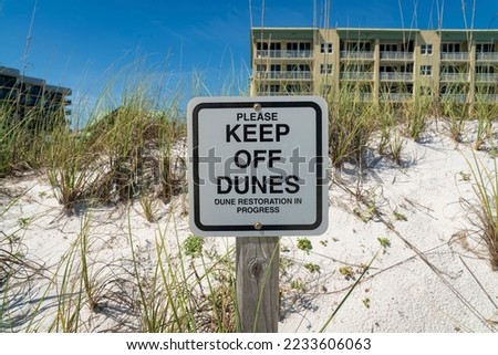 Please Keep Off Dunes, Dune Restoration in Progress signage on white sand with grass at Destin, FL. Close-up of a sign with multi-storey building with balconies against the clear sky at the back.