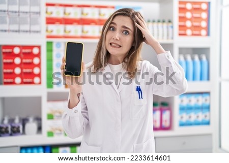 Blonde caucasian woman working at pharmacy drugstore showing smartphone screen stressed and frustrated with hand on head, surprised and angry face  Royalty-Free Stock Photo #2233614601