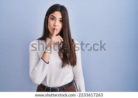 Young brunette woman standing over blue background asking to be quiet with finger on lips. silence and secret concept. 