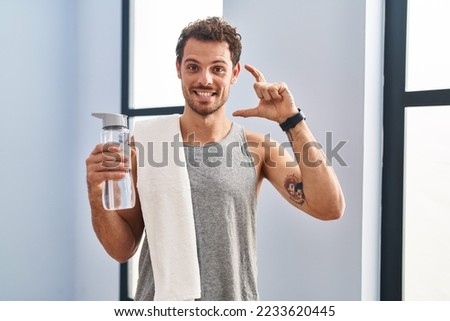 Young hispanic man wearing sportswear drinking water smiling and confident gesturing with hand doing small size sign with fingers looking and the camera. measure concept.  Royalty-Free Stock Photo #2233620445